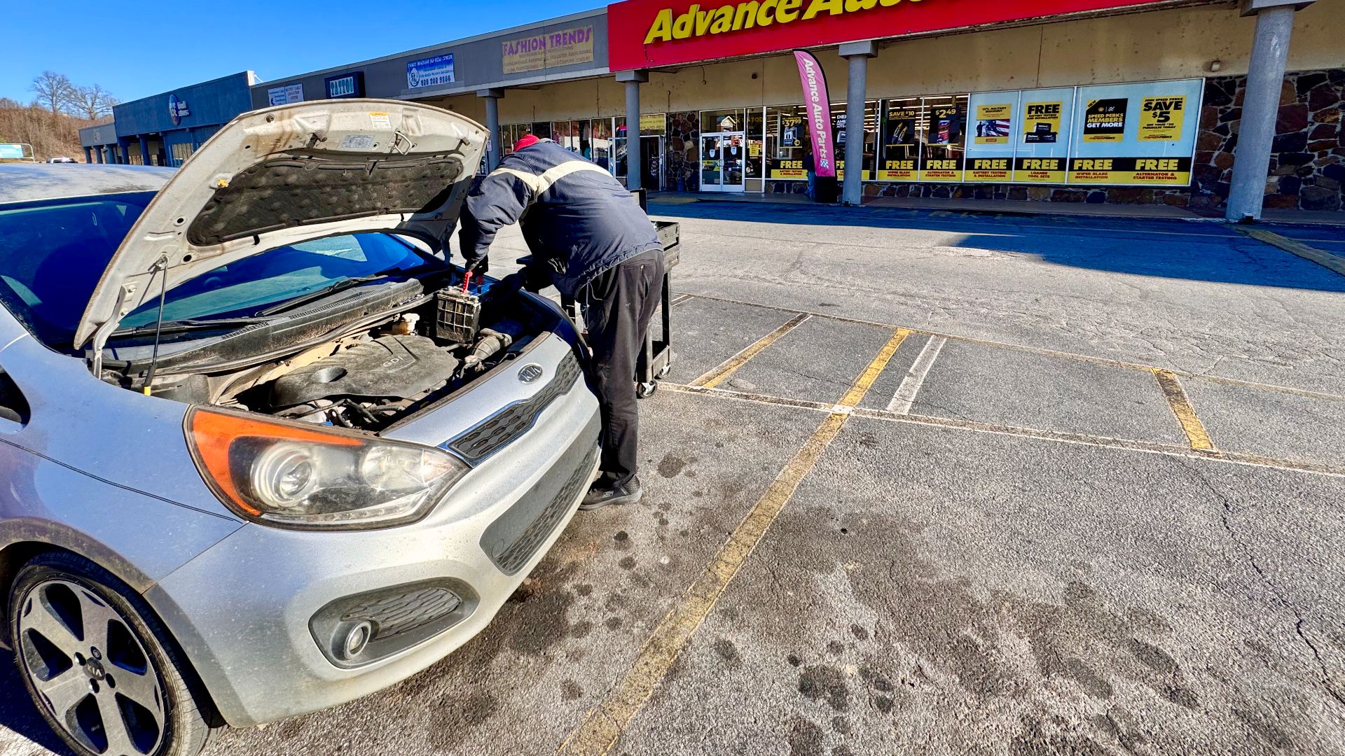 An Advance Auto Parts store employee changes a car battery in a parking lot in front of the shop