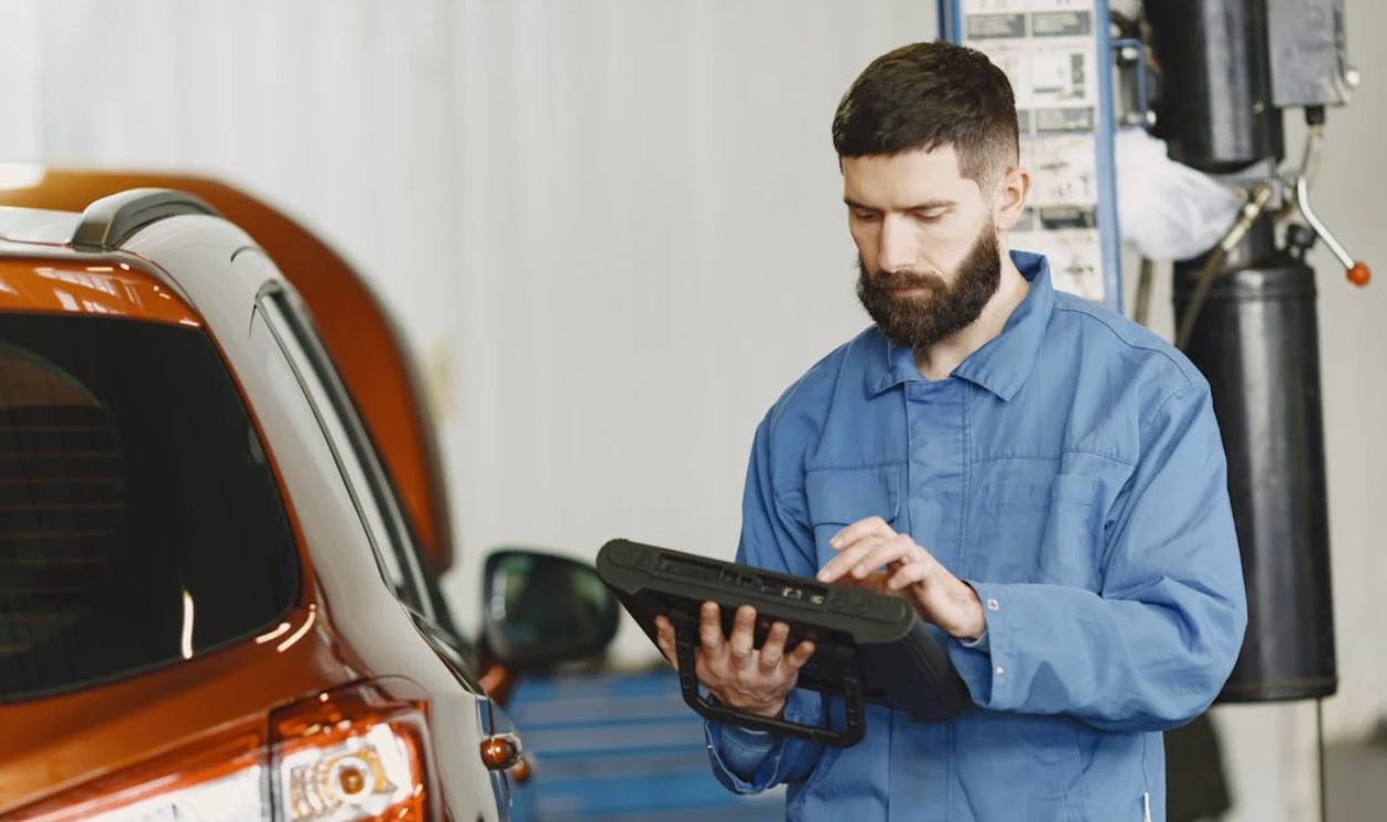 Man in Blue Coveralls Standing Beside an Orange Car Using an Automotive Diagnostic Tool