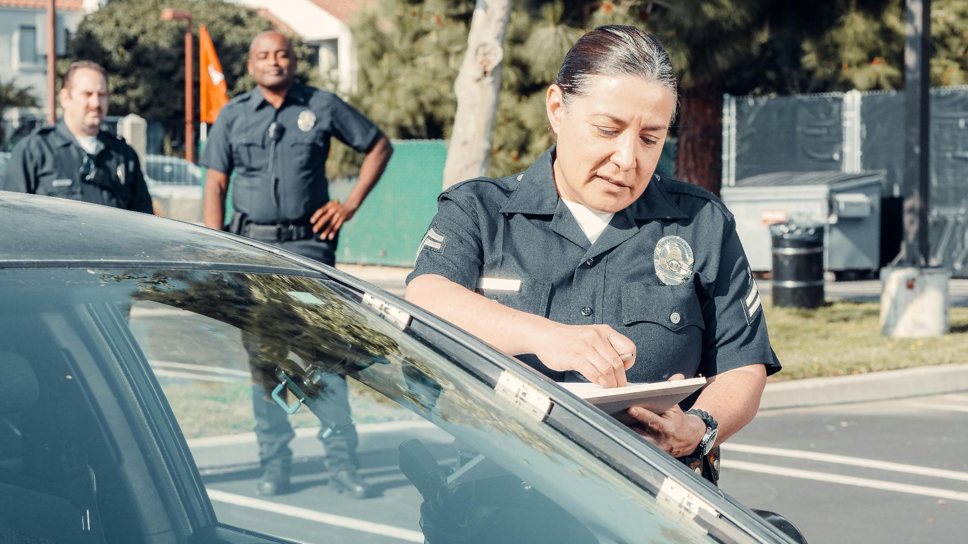 Police officers handling a traffic violation in a parking lot, focus on policewoman writing a ticket.