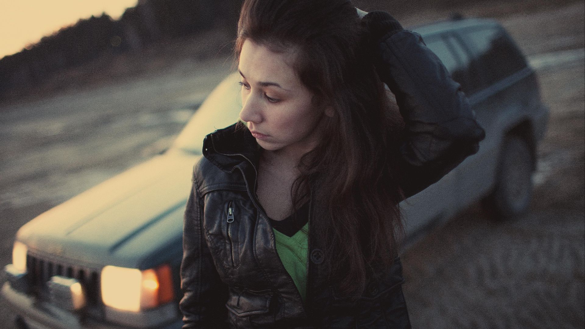 A thoughtful woman in a leather jacket stands near a car in an outdoor setting during dusk.
