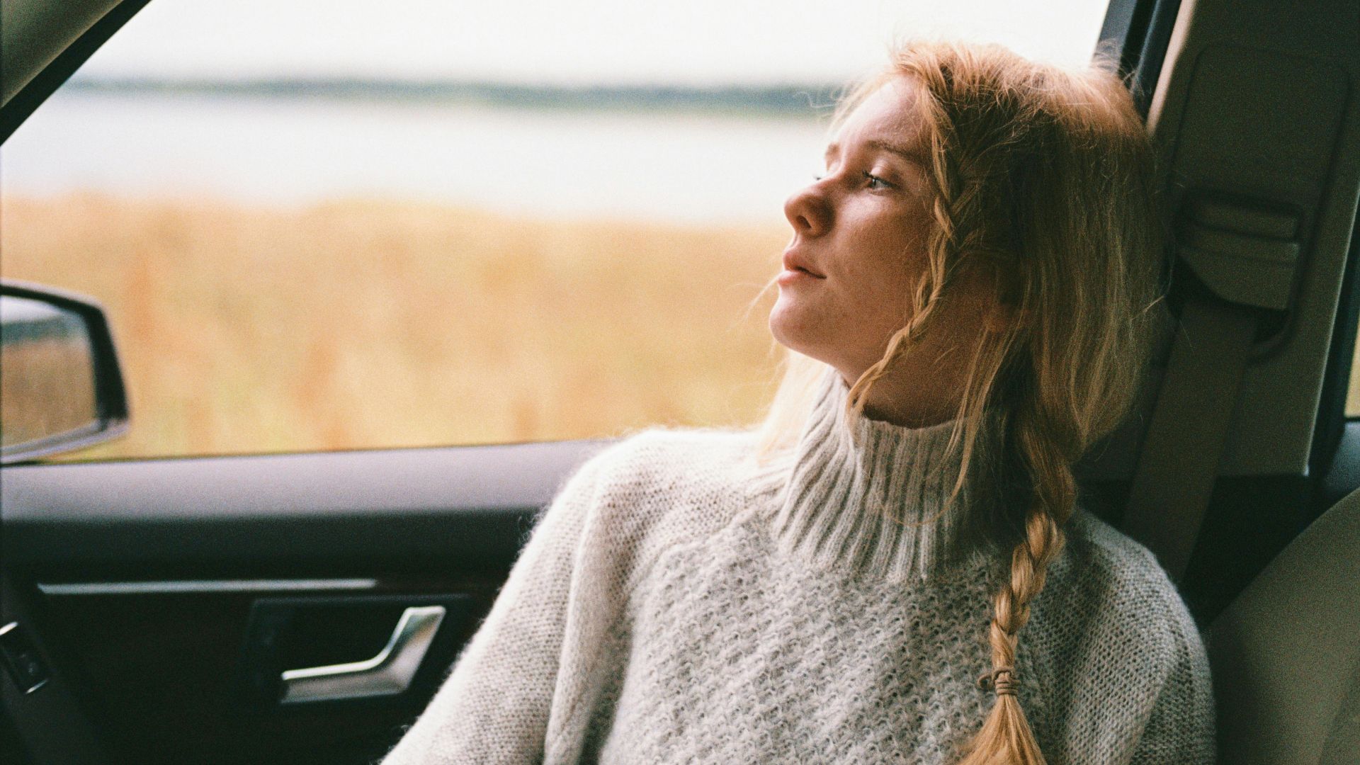 A thoughtful woman with braided hair gazes out a car window during a daytime road trip.