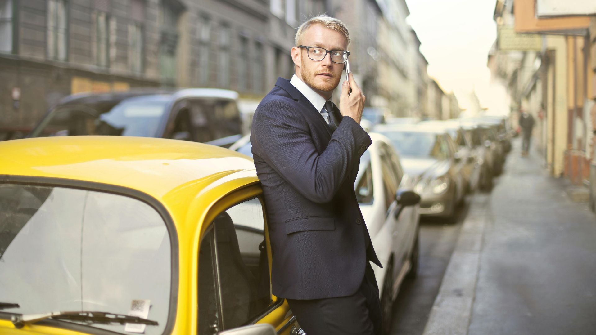 Businessman in a suit leaning on a yellow car making a phone call on a city street.