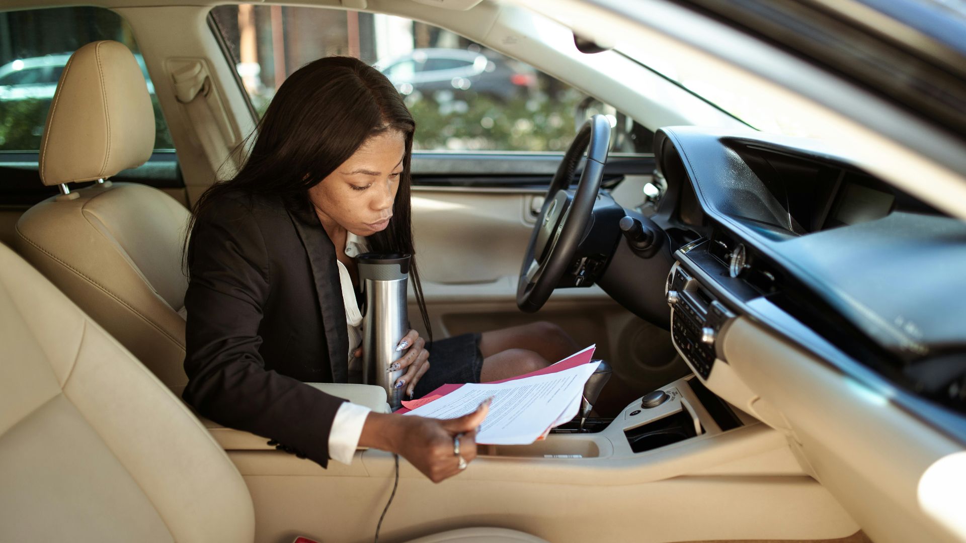 A focused businesswoman reads documents in her car, multitasking with a tumbler.