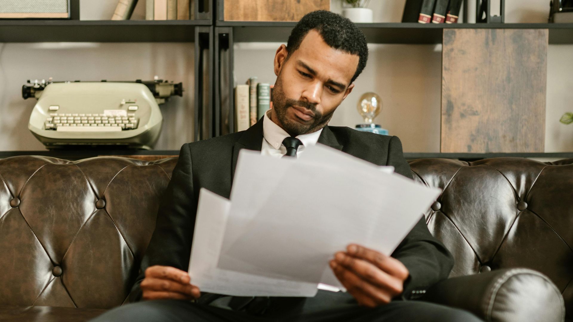 Adult man in a suit reading paperwork on a leather couch in a stylish office setting.