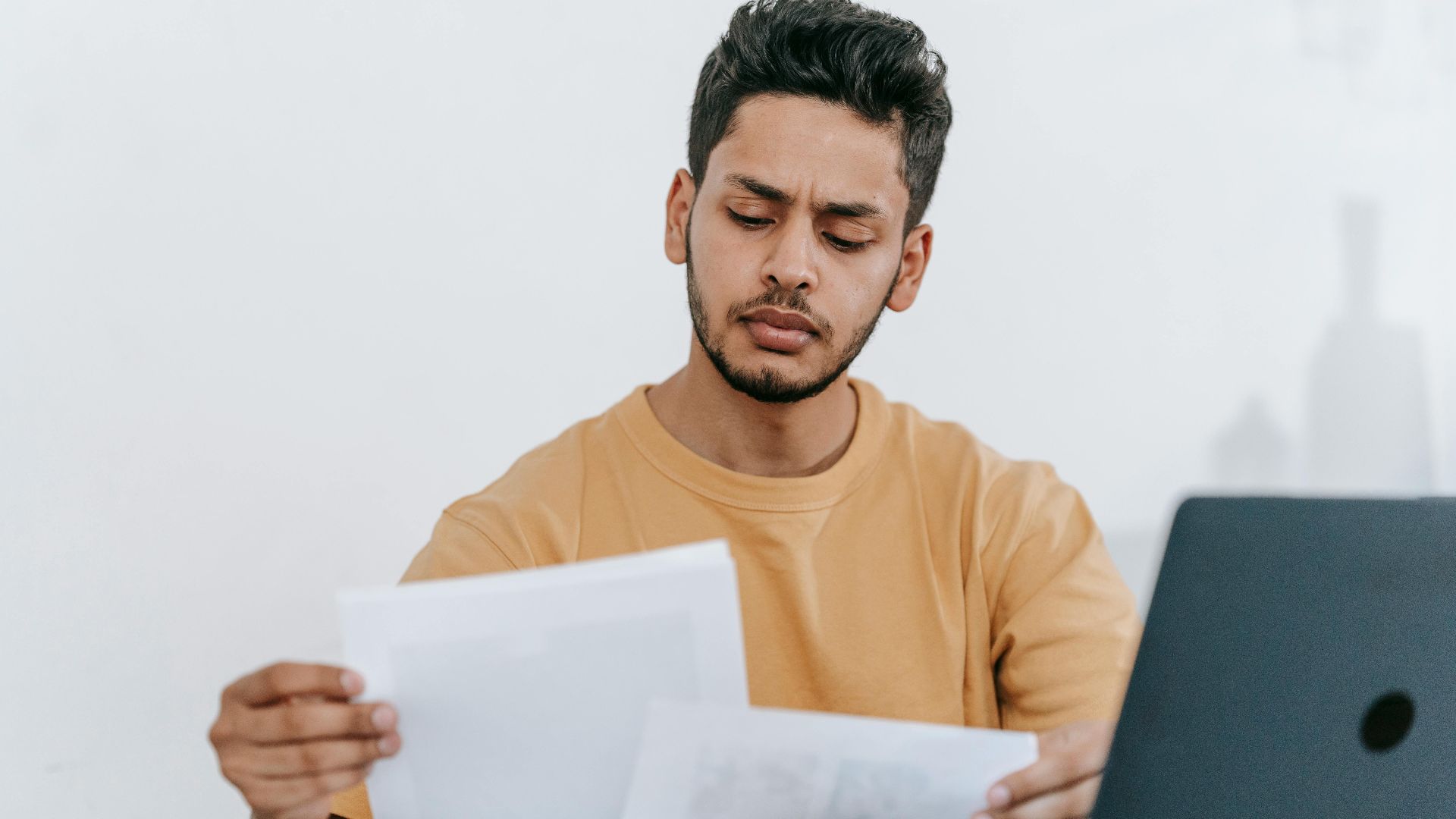 Focused young man reviewing paperwork at his desk, showcasing a business setting with a laptop indoors.