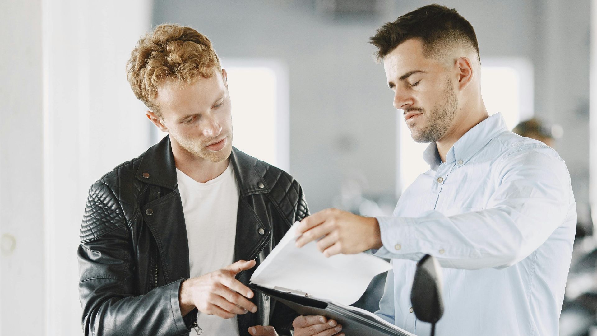 Two men in a motorcycle dealership discussing details over documents next to a bike.
