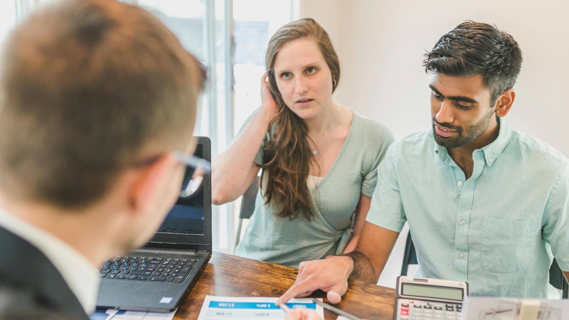 A young couple consults with a realtor about buying a home, indoors in a modern office setting.
