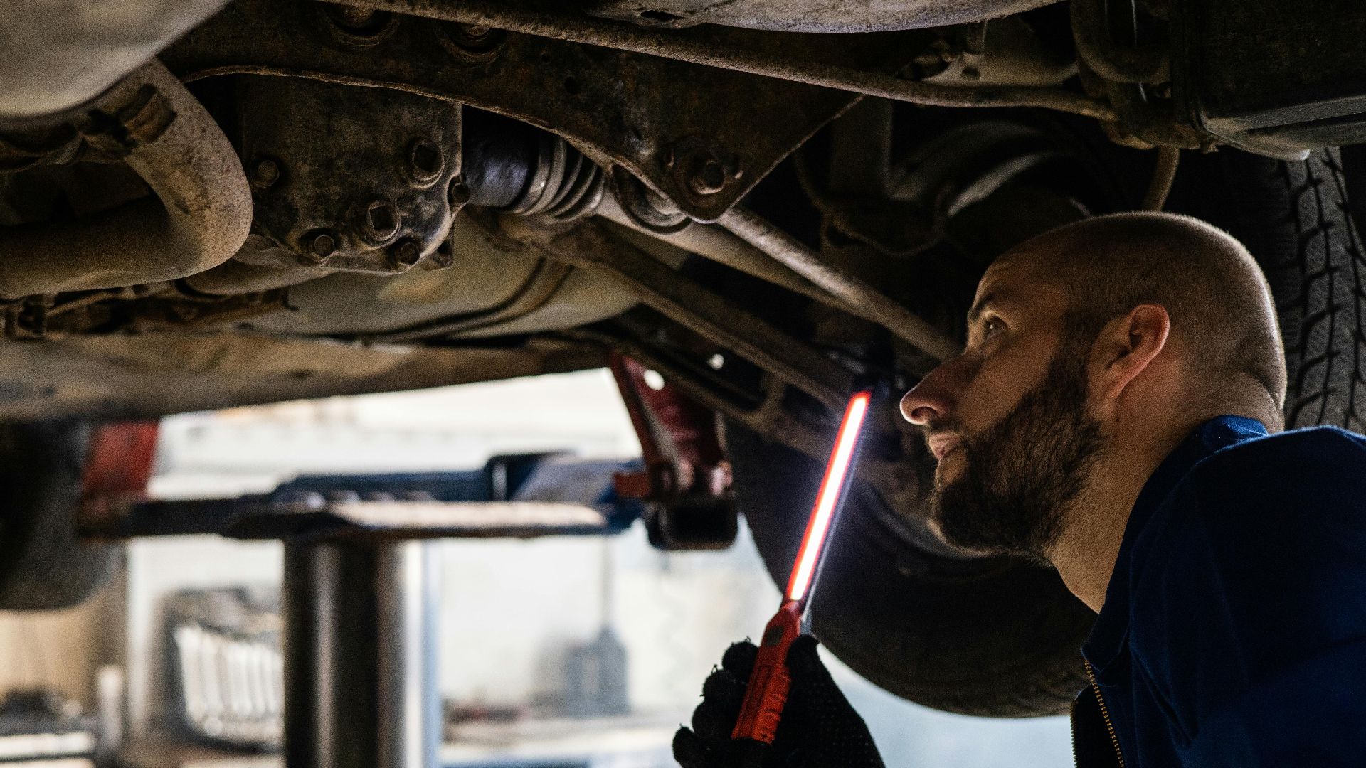 Mechanic using light to inspect vehicle undercarriage in workshop for maintenance.