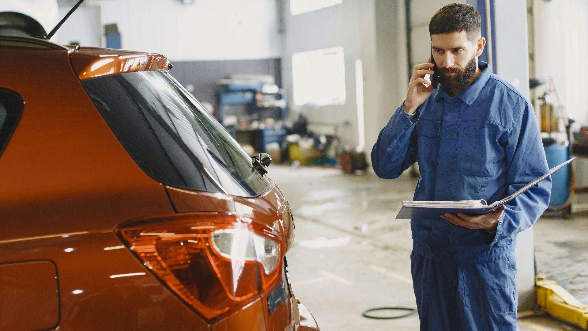 Mechanic in a blue uniform talks on phone beside a red car in an auto repair shop.