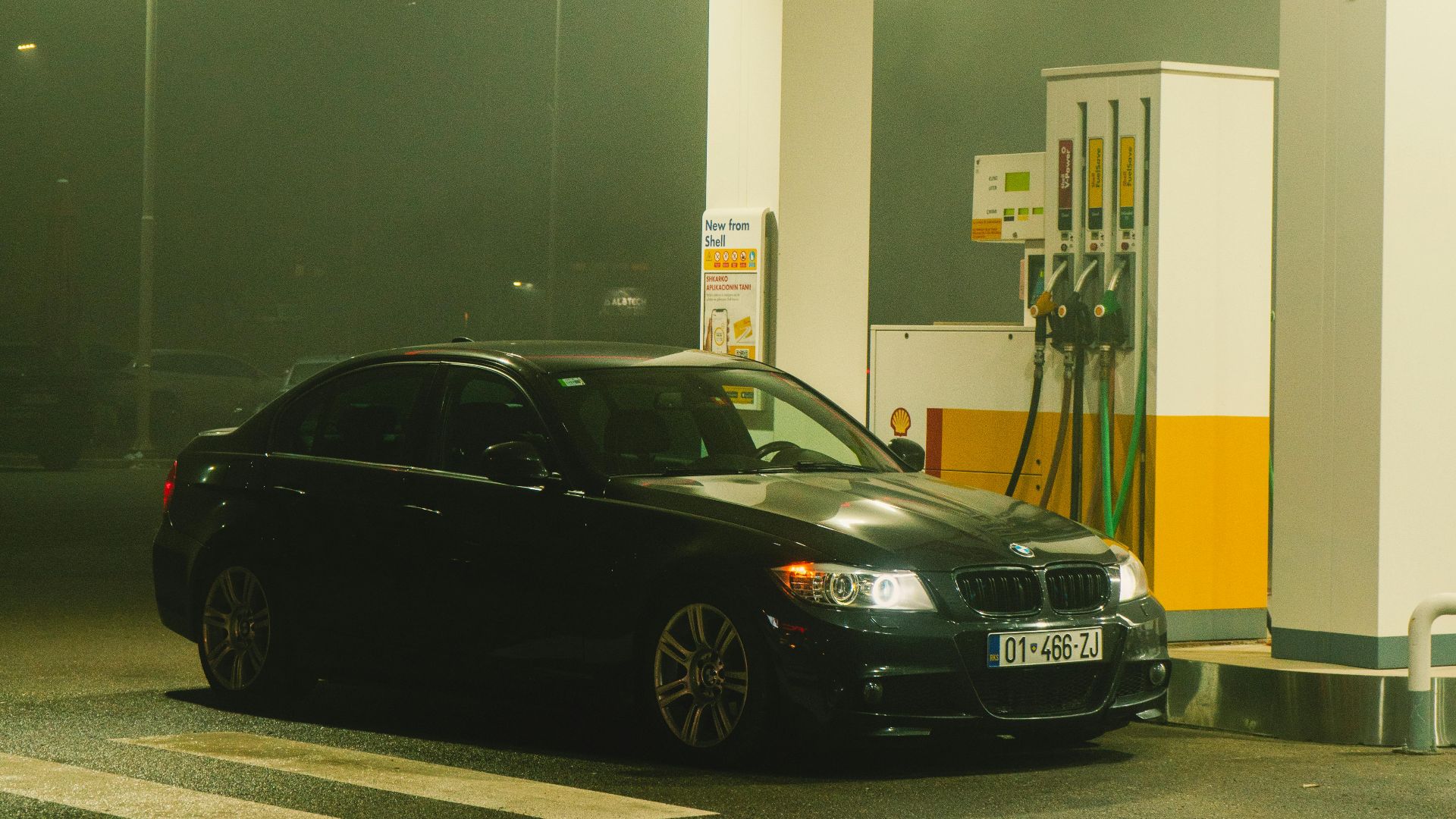 BMW sports car refueling at a Shell gas station under night lights in a misty urban setting.