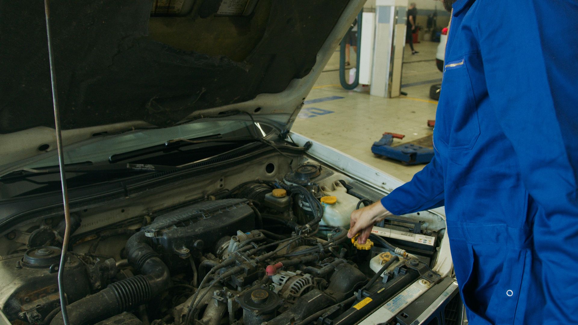 Mechanic in blue coveralls inspecting an open car engine in an automotive workshop.