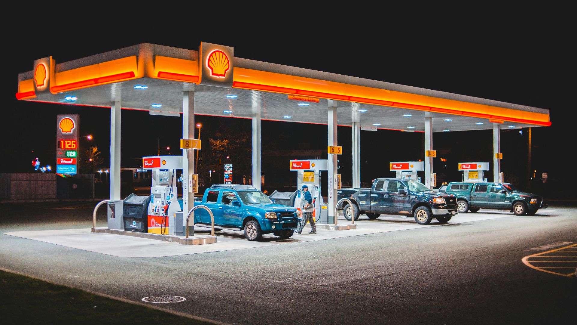A glowing Shell gas station at night with parked vehicles and vibrant lighting.