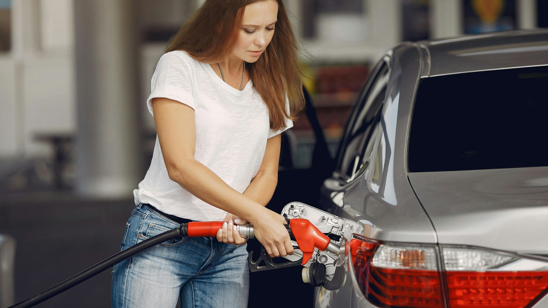 Attentive female driver in casual outfit and headband filling up modern automobile with automotive fuel gun on petrol station while looking down