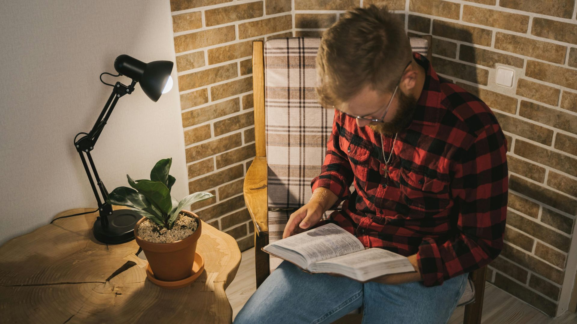Man in plaid shirt reading in a cozy, well-lit corner with a plant and table lamp.