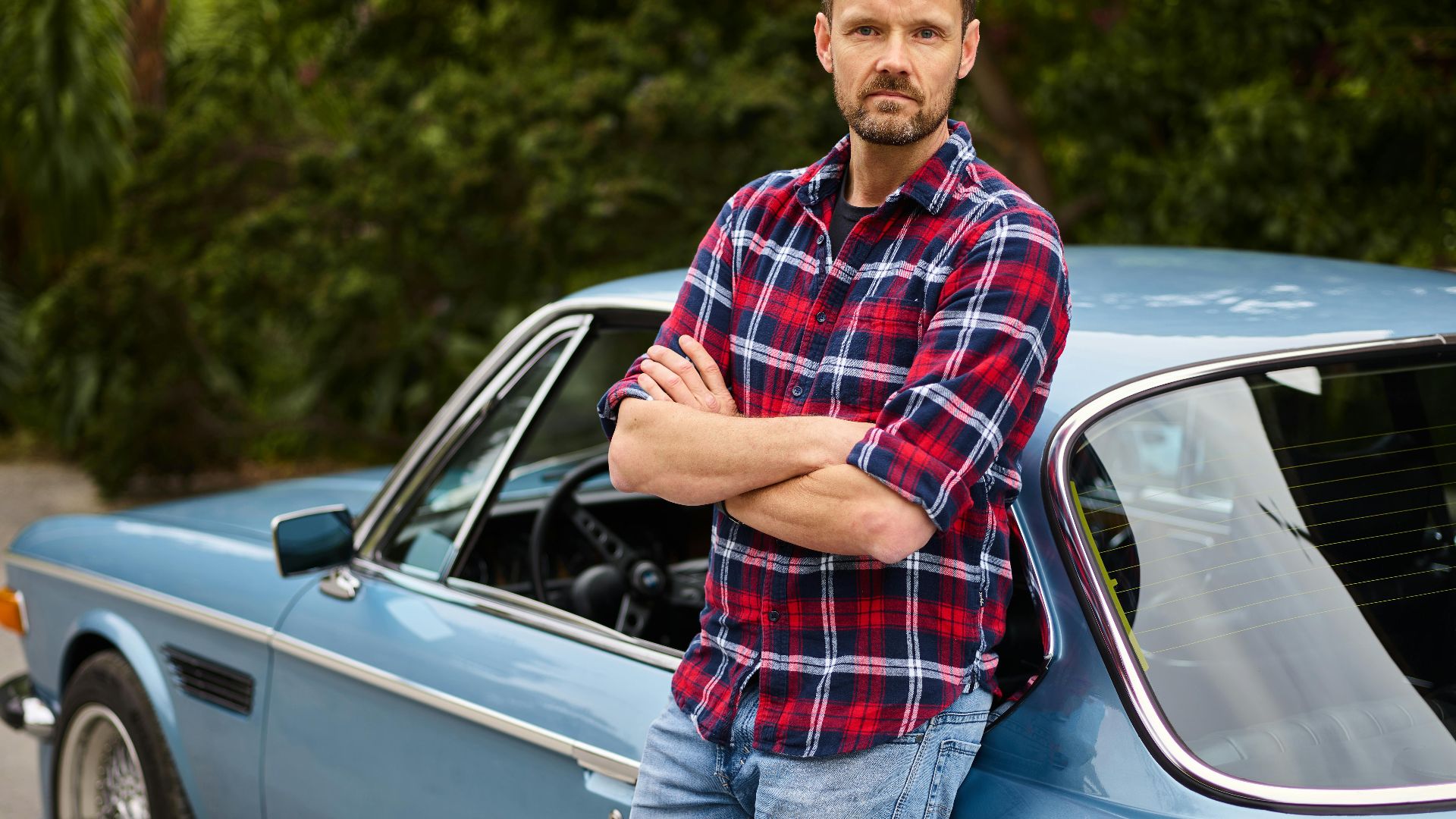 Man with arms crossed leaning on a vintage car in a serene countryside setting.