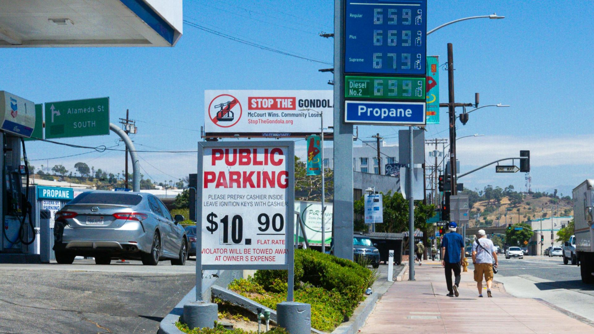 People walking near a Chevron gas station with prominent signage and parked vehicles on a sunny day.