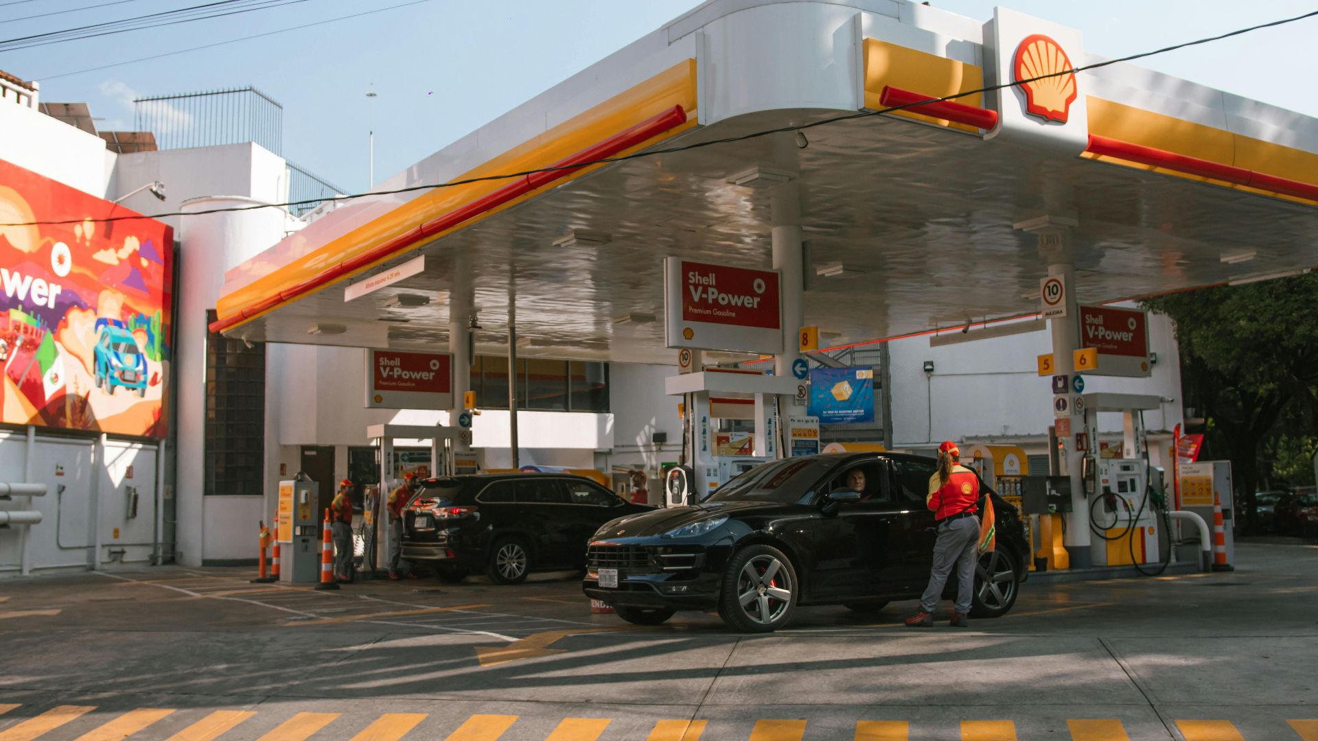 A Shell gas station with vehicles refueling and attendants assisting drivers on a sunny day.