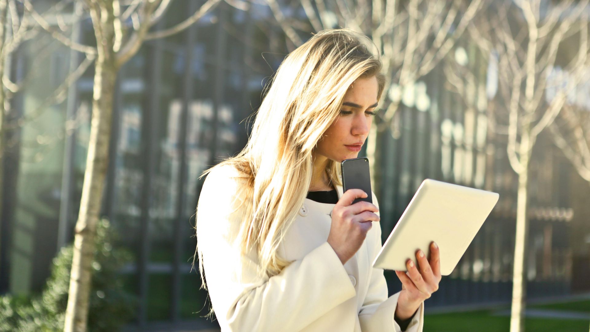 A young woman using her tablet and smartphone outdoors in a city park.