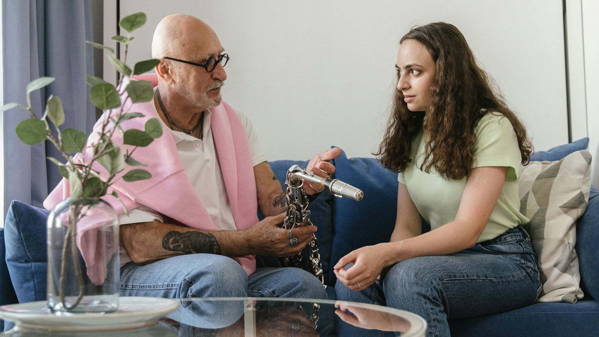 Elderly man and young woman seated on sofa discussing saxophone indoors.