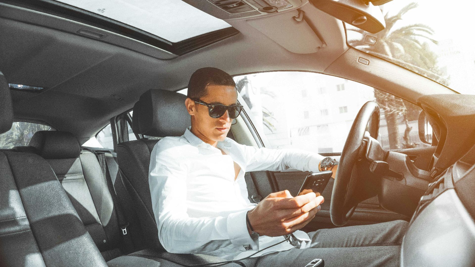A man wearing sunglasses texts on his phone while driving a car in Morocco.