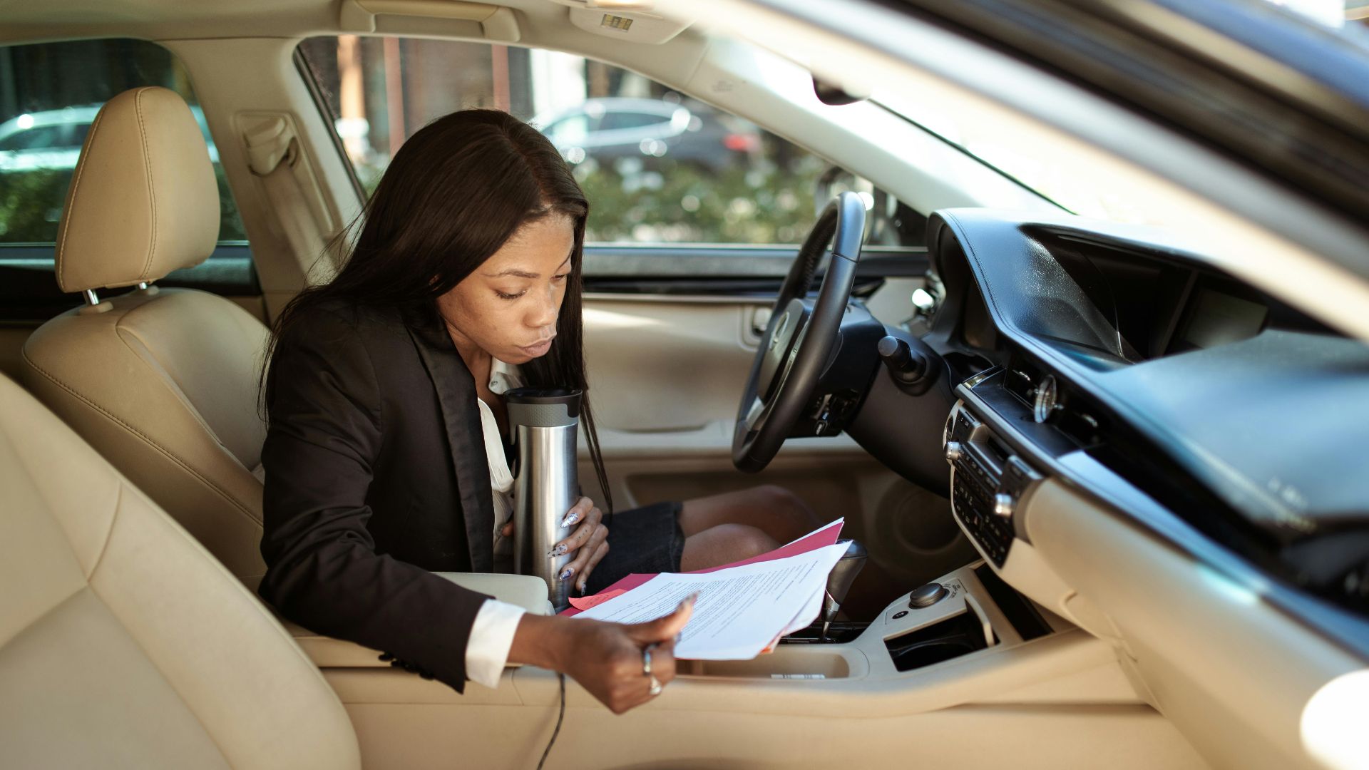 A focused businesswoman reads documents in her car, multitasking with a tumbler.