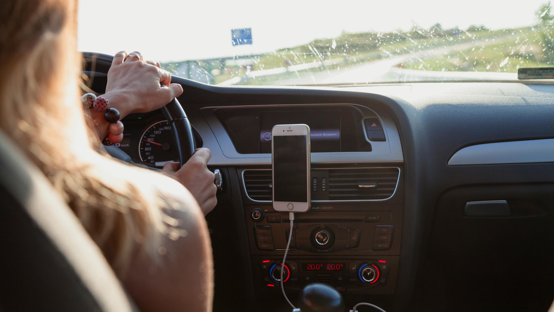 View from behind a woman driving a car on a sunny day with a phone mounted on the dashboard.