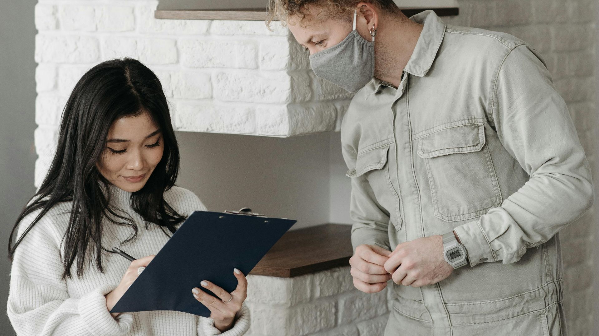 An Asian woman signs a clipboard for a delivery man wearing a face mask indoors.