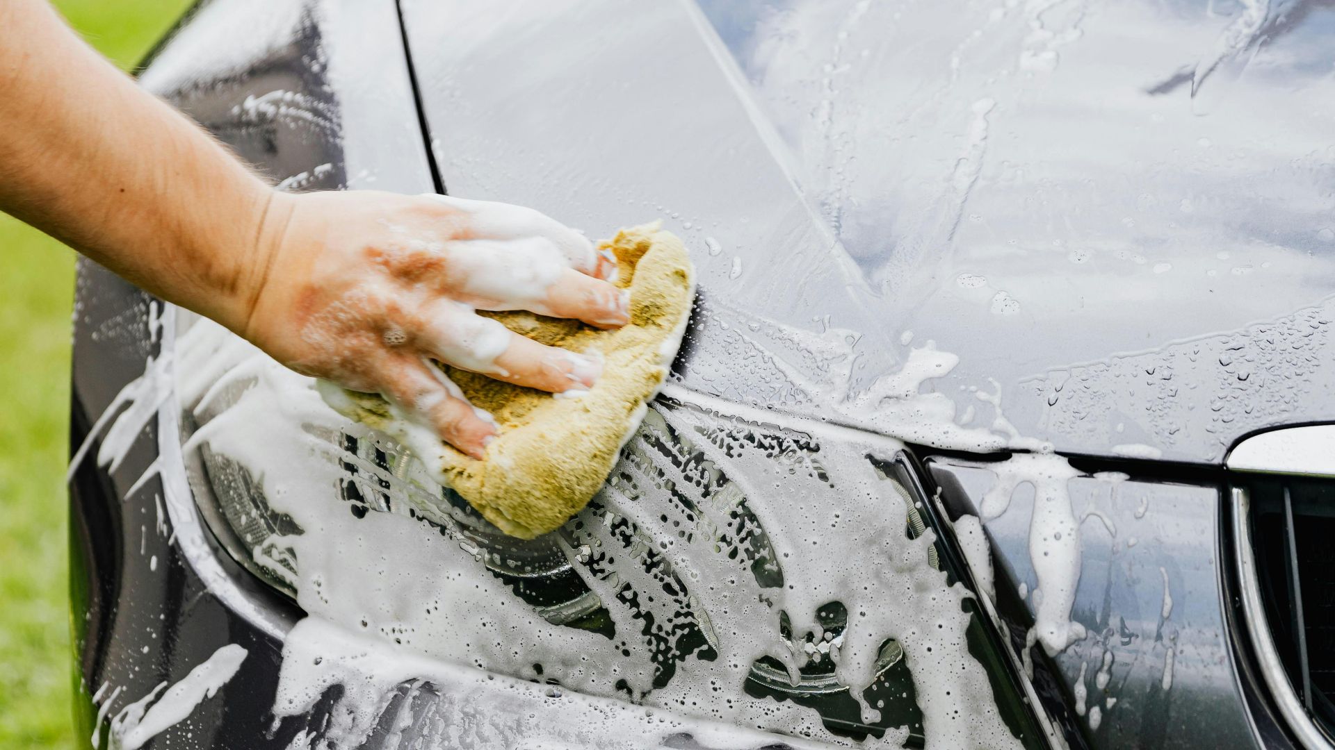 Person washing a black car's headlight with a sponge and soap outdoors.