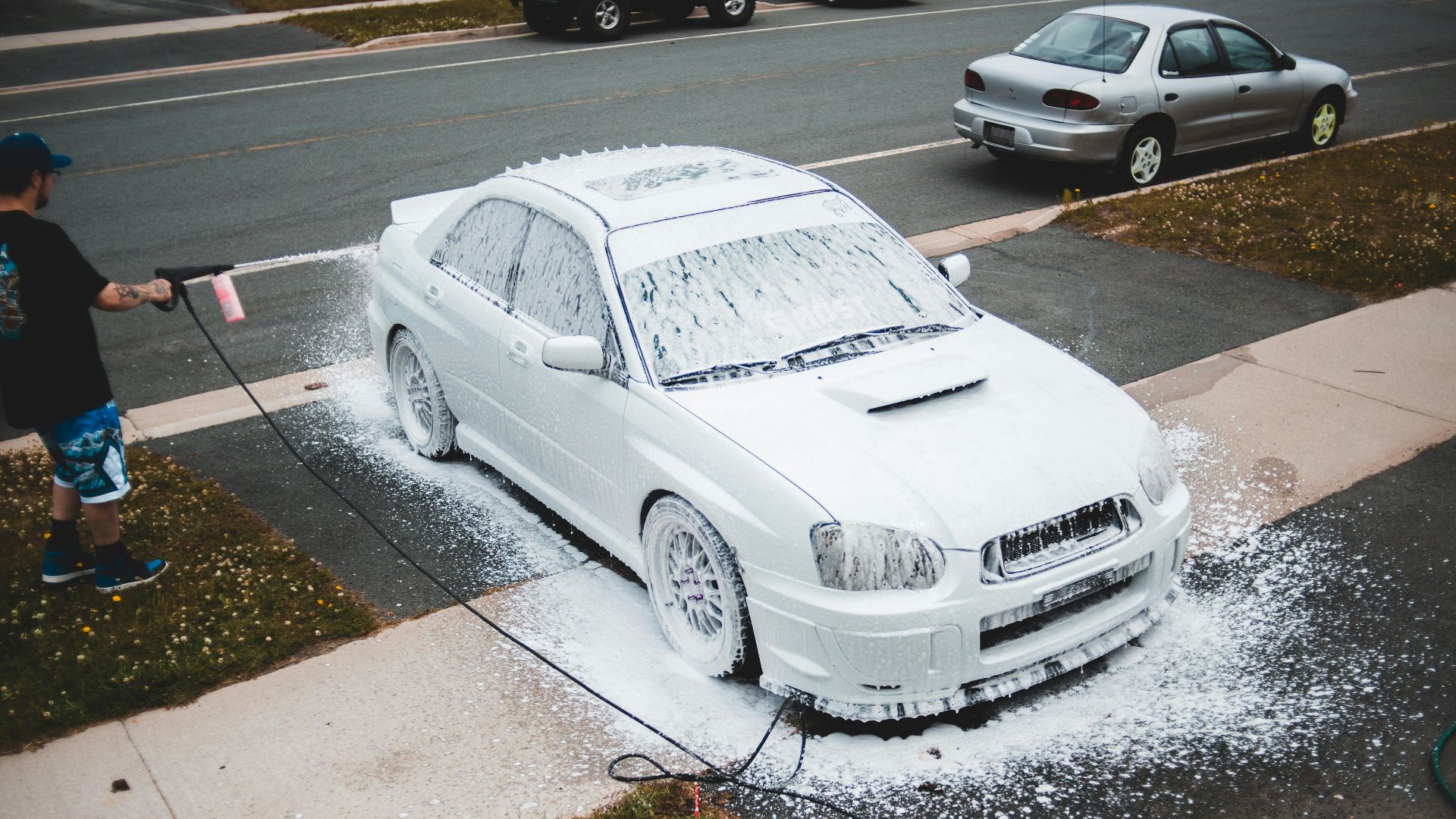 A man using a pressure washer to clean a white car with foam on a suburban driveway.
