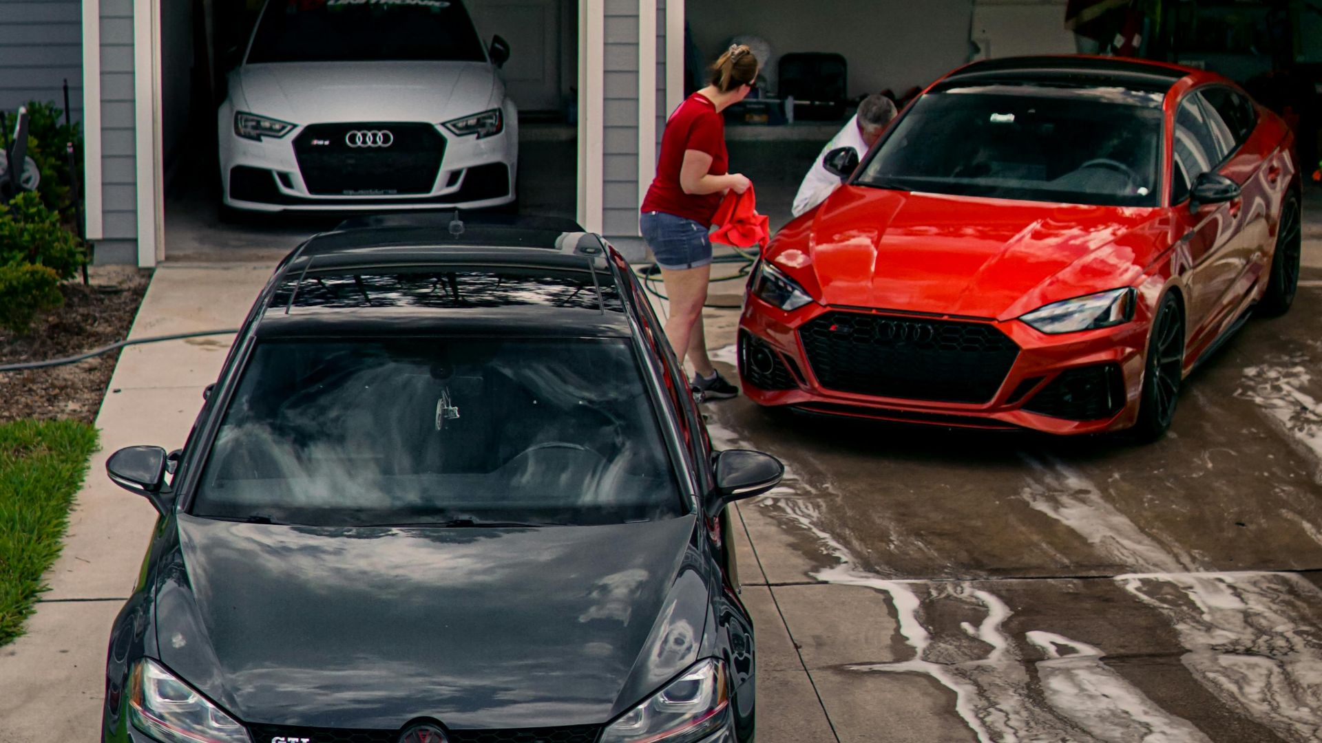 A person washing modern cars in a driveway surrounded by a lush backyard.