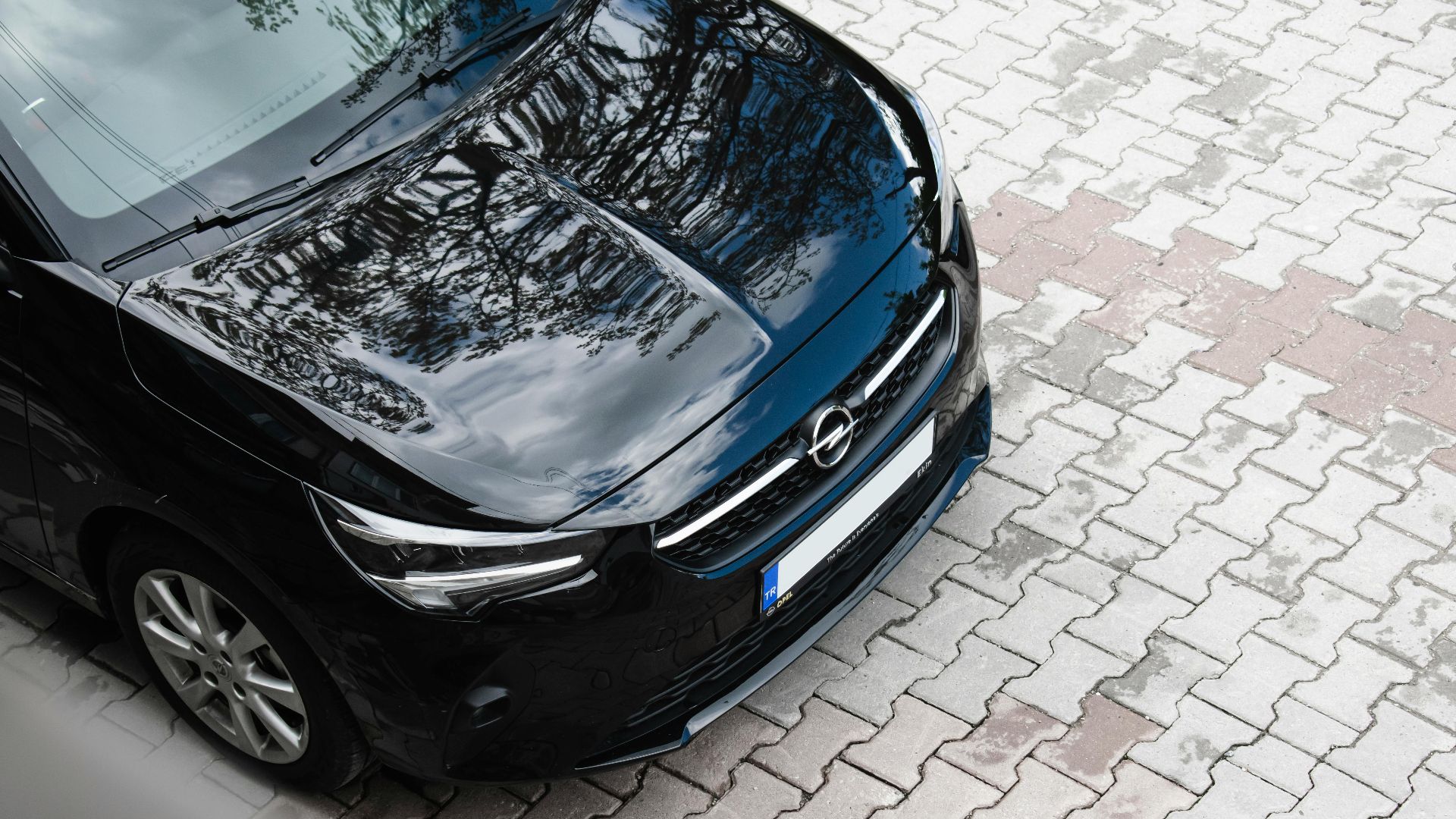 High angle view of a black Opel Corsa reflecting trees, parked on a patterned pavement.