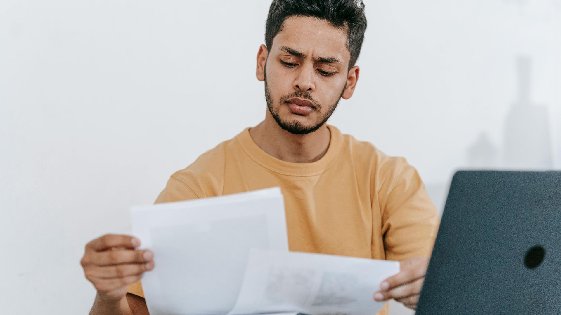 Focused young man reviewing paperwork at his desk, showcasing a business setting with a laptop indoors.