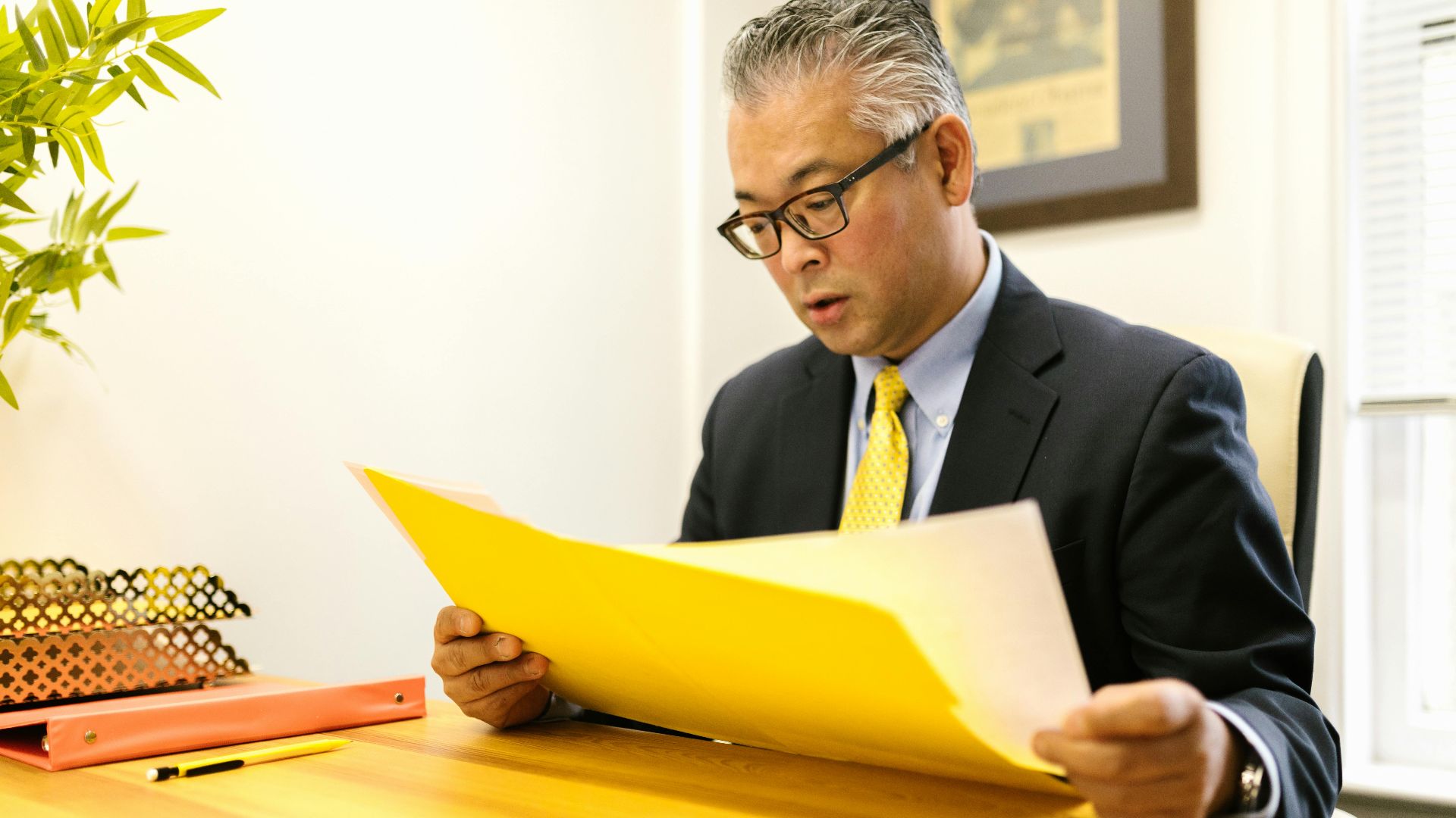 Asian businessman in corporate attire reading documents at office desk with a yellow folder.
