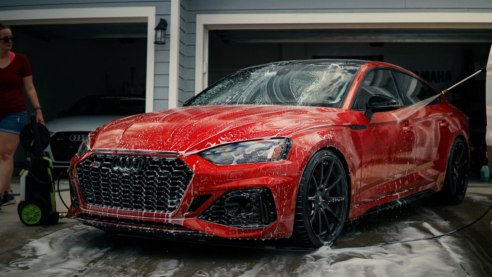 A red sports car is being cleaned with soap in a driveway, featuring a modern garage background.
