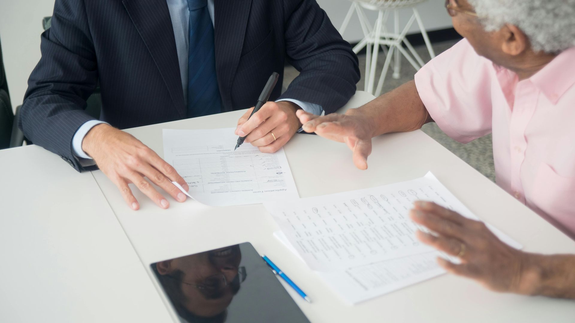 A business meeting between an elderly client and a consultant discussing documents at an office table.