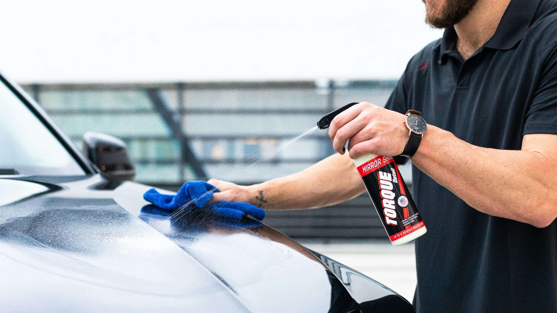 Close-up of a man cleaning a car hood with spray and microfiber cloth outdoors.