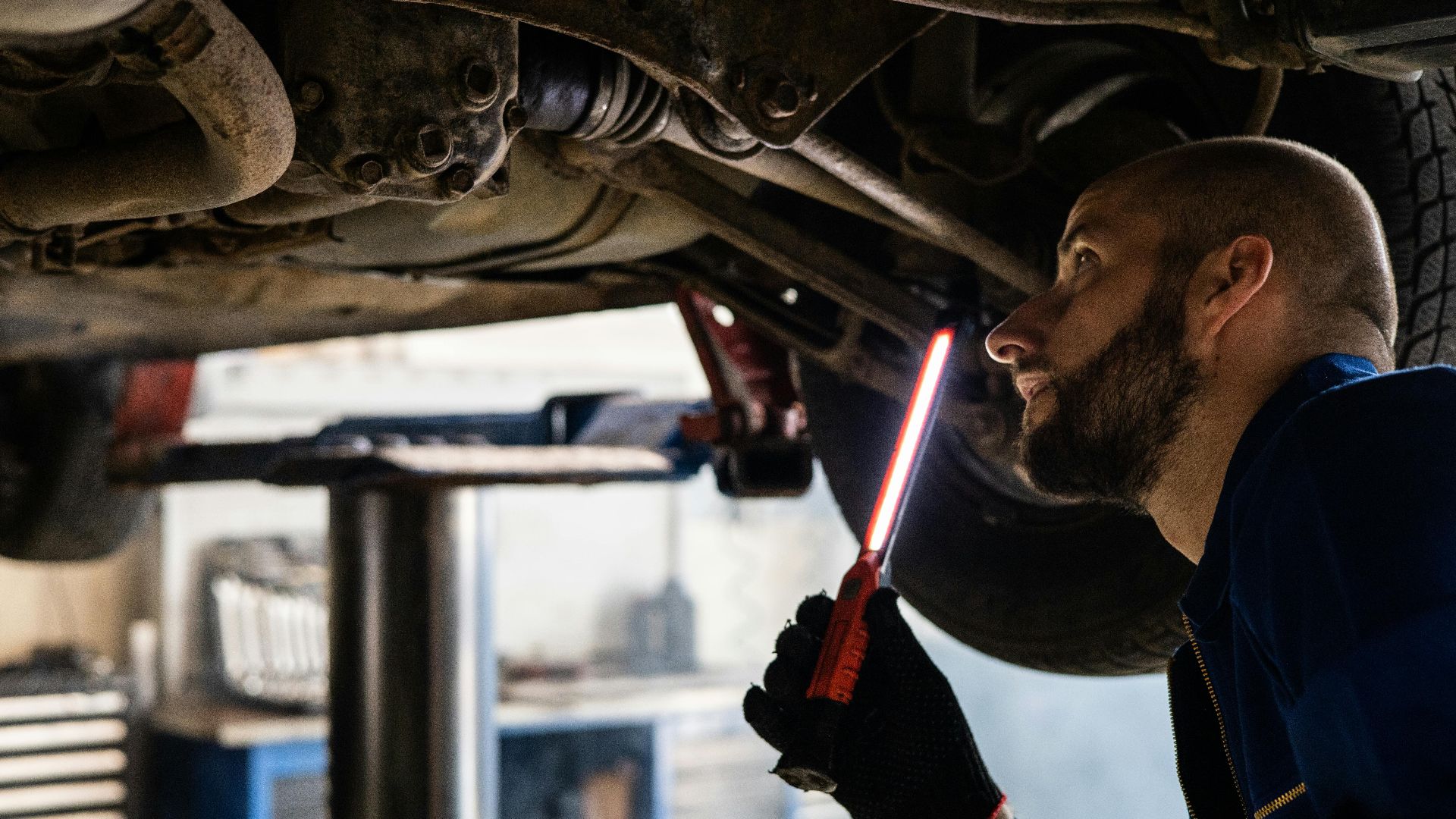 Mechanic using light to inspect vehicle undercarriage in workshop for maintenance.
