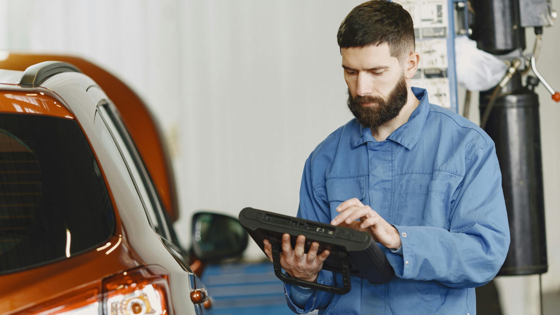 A mechanic in coveralls using a diagnostic tool at an auto repair shop beside a car.