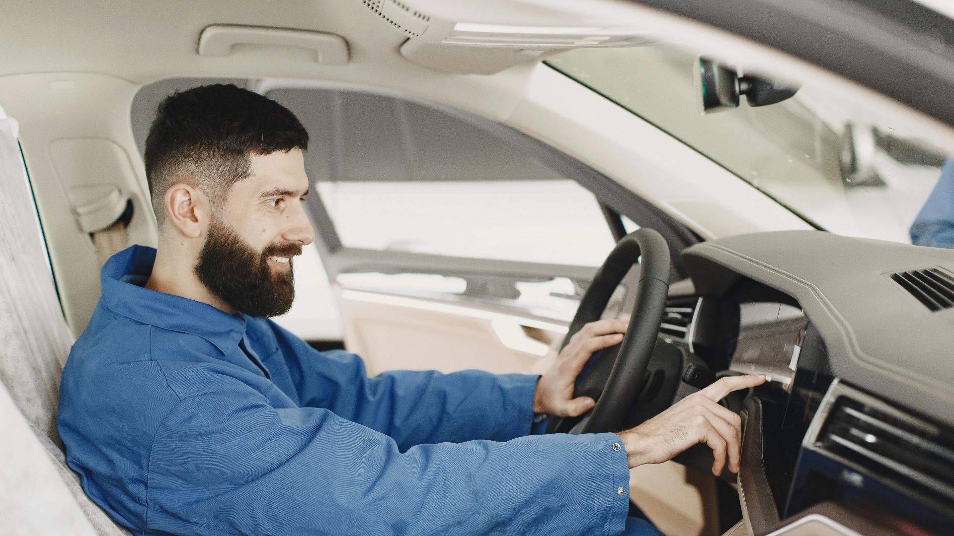 Mechanic in blue coveralls interacts with car dashboard, smiling and focused.