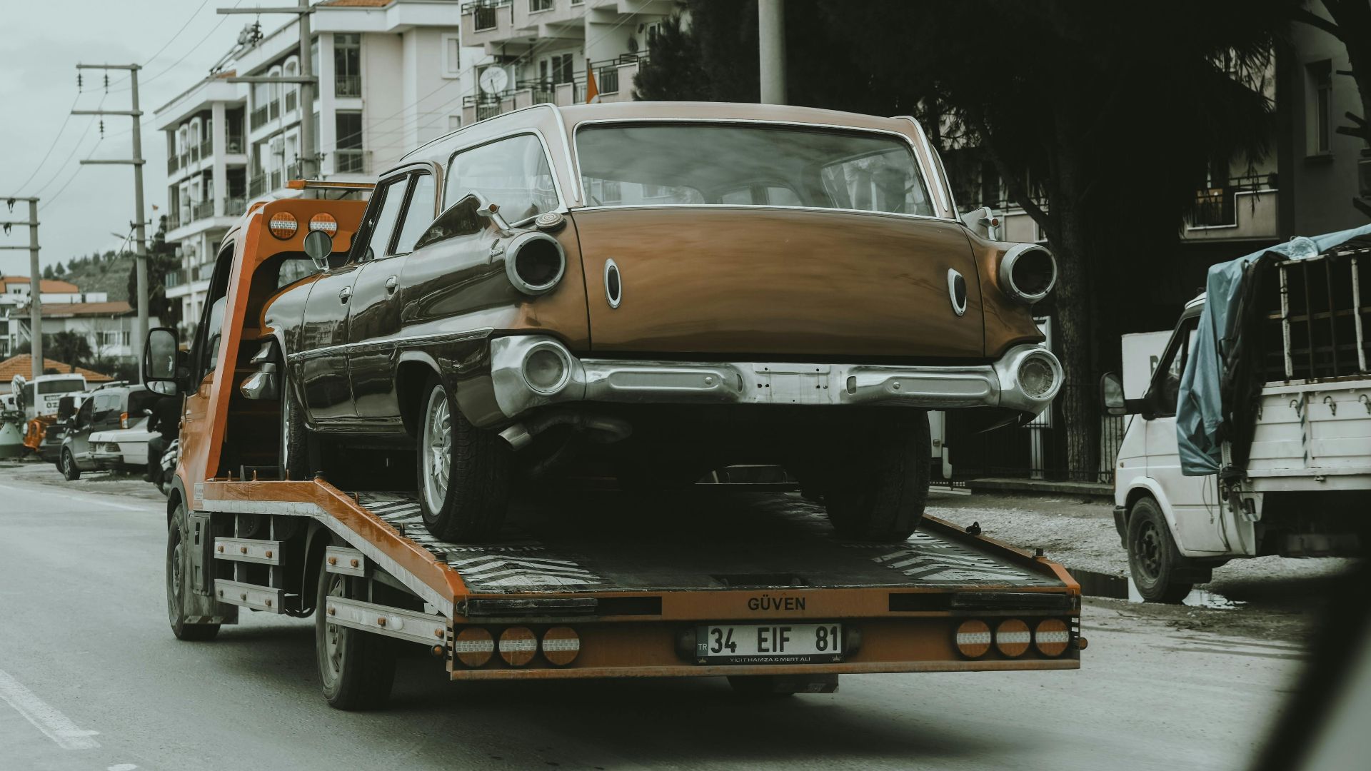 A vintage car loaded on a tow truck in a city street, surrounded by modern buildings.