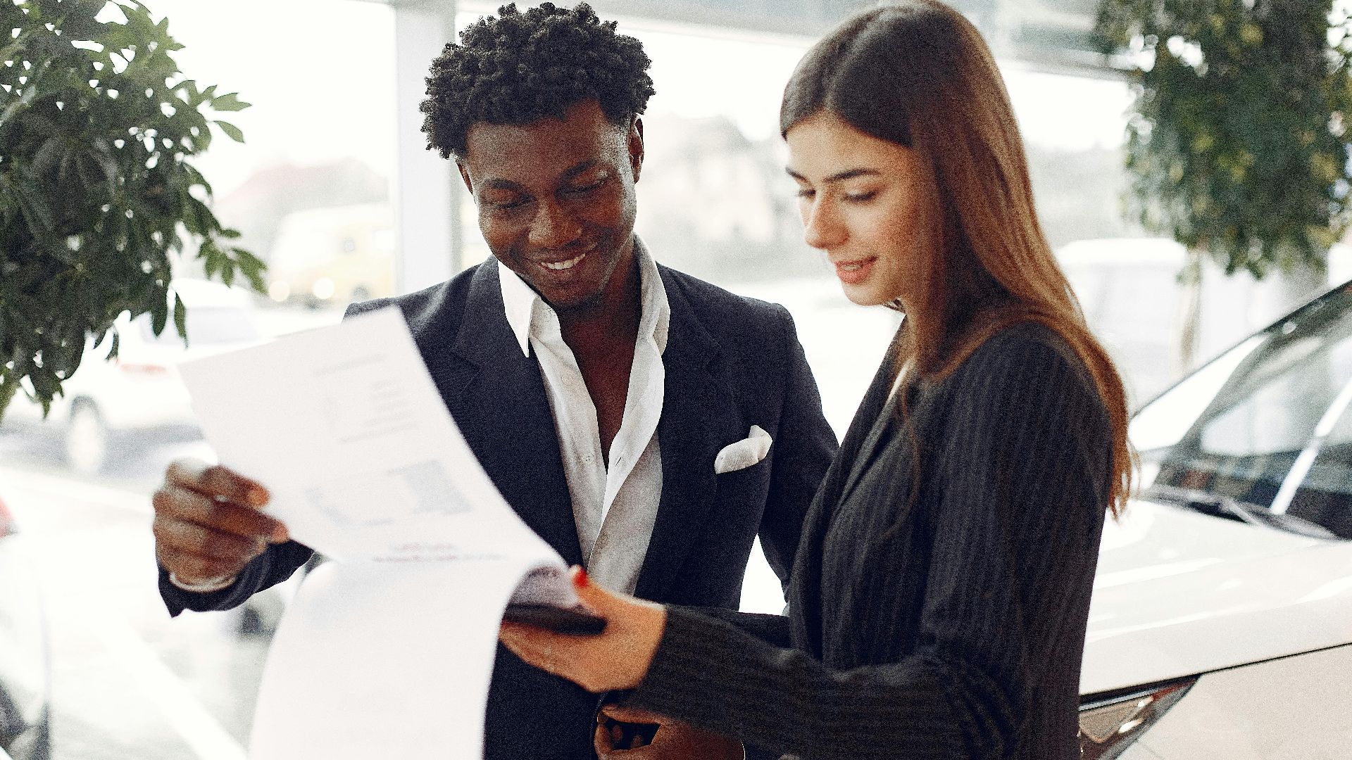 Cheerful smiling multiethnic businessman in classy suit and professional elegant female consultant standing close and reading contract details in car showroom