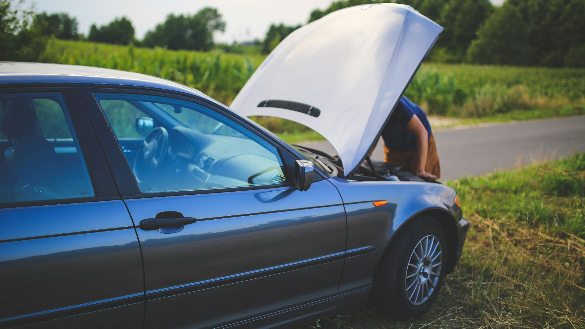 Man checking car engine with hood open by the side of a rural road.