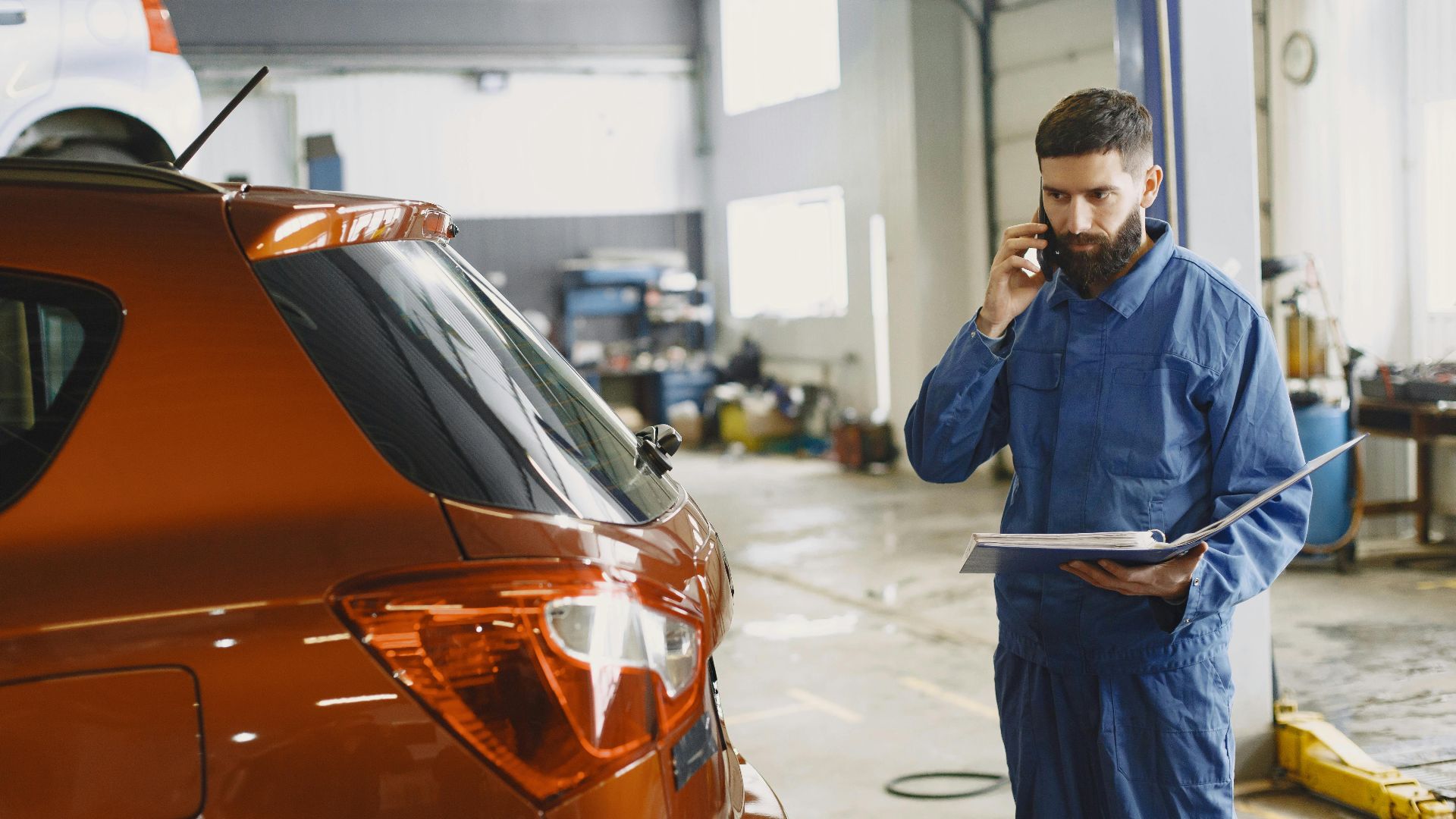 Mechanic in a blue uniform talks on phone beside a red car in an auto repair shop.