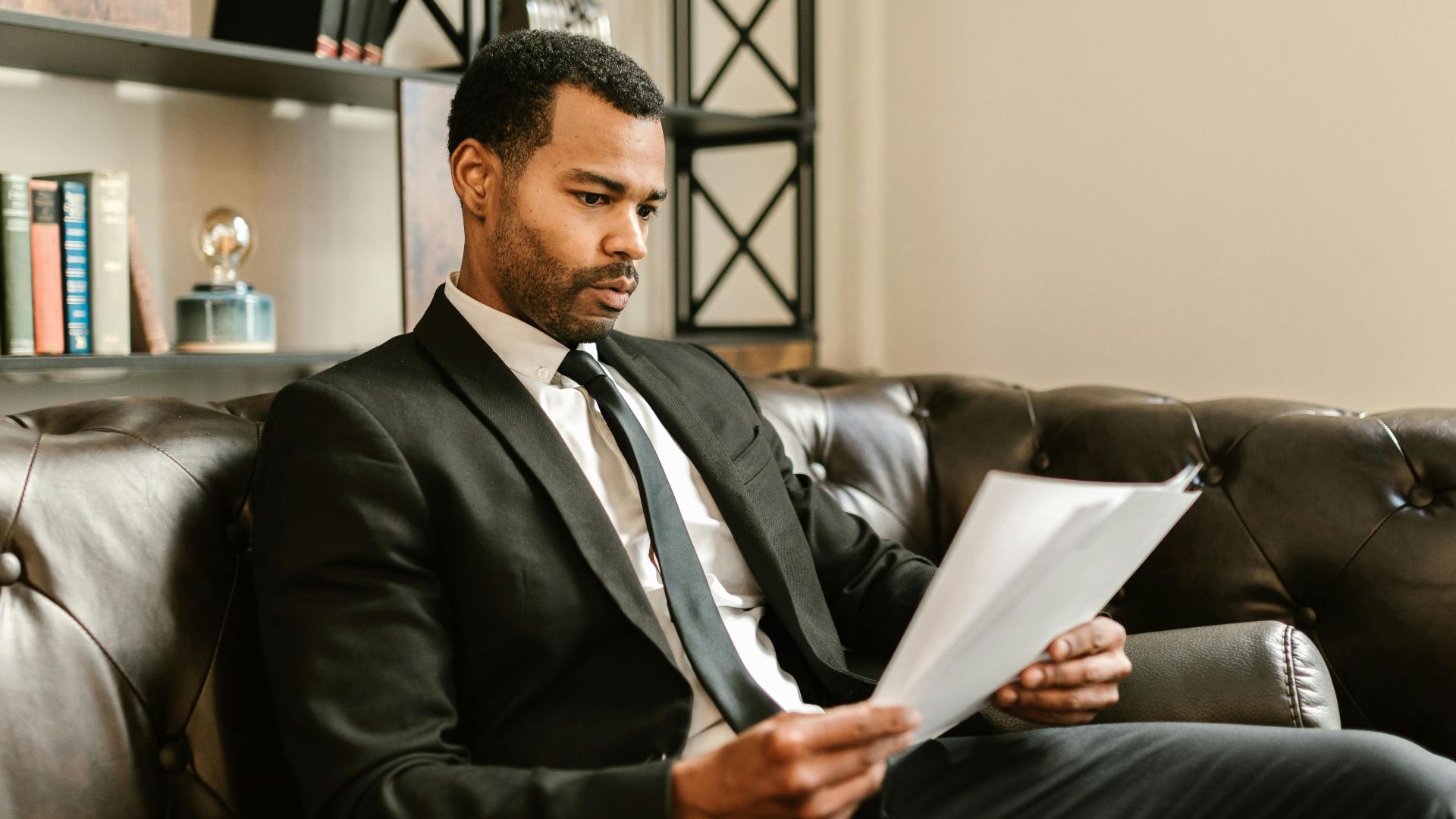 Businessman in suit reading papers on a leather sofa in a modern office setting.