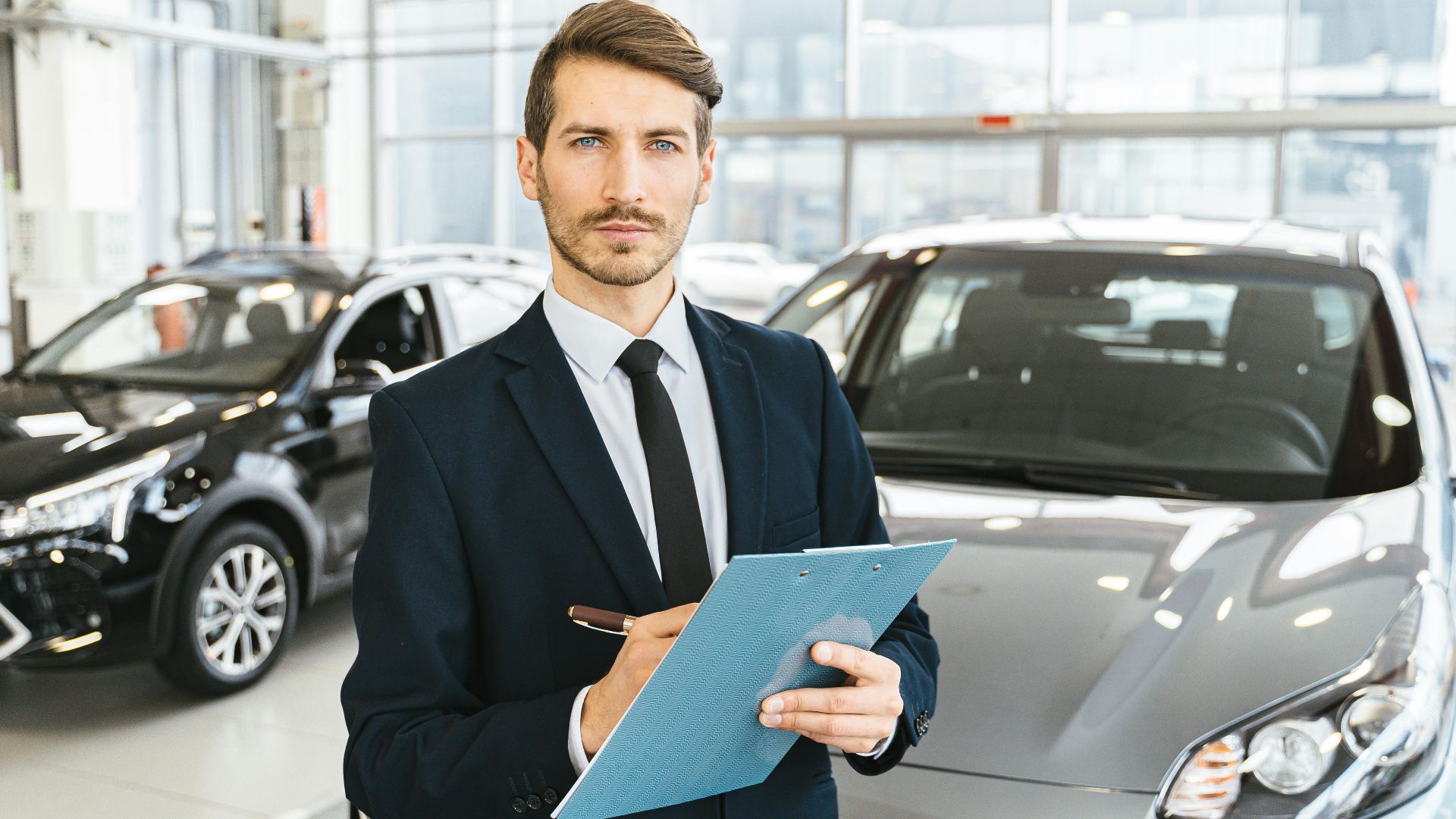 A confident car salesman in a showroom, holding a clipboard and pen.