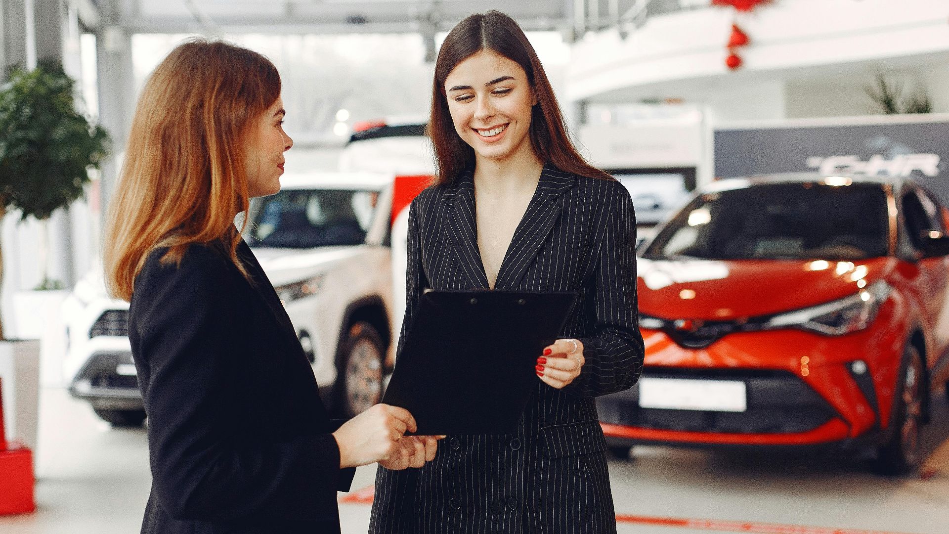 Cheerful young friendly dealer in formal stylish black dress showing contract to smiling female customer in black jacket while standing in car showroom against new shiny automobiles