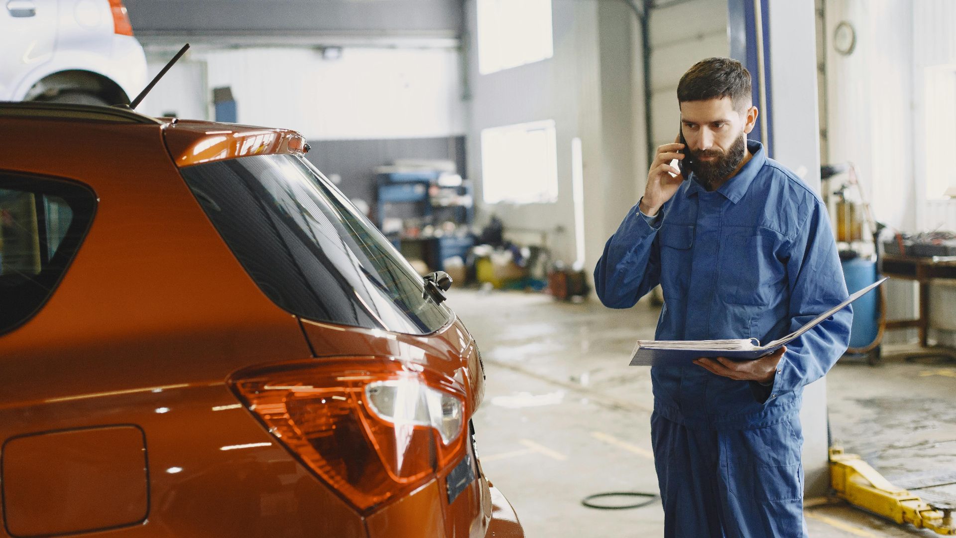 Mechanic in a blue uniform talks on phone beside a red car in an auto repair shop.