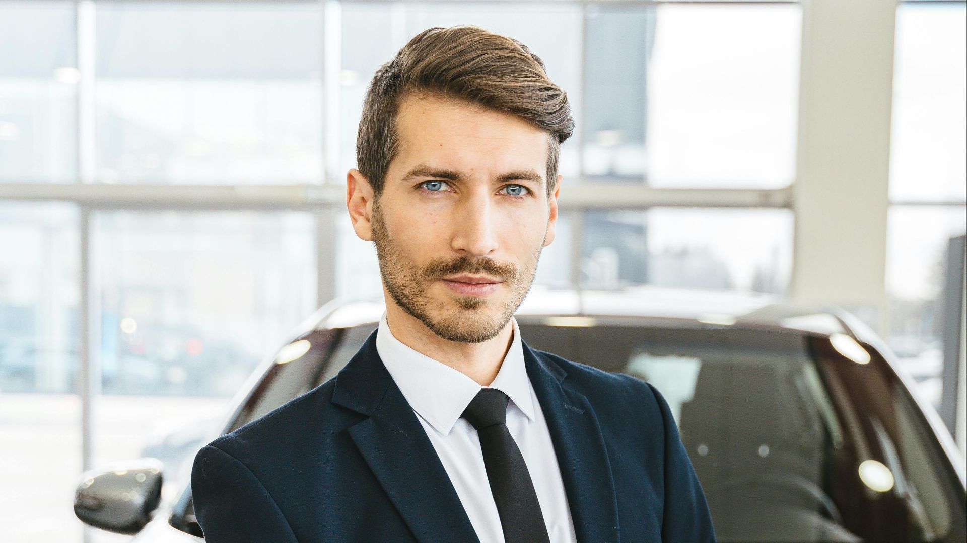 Sales professional in a suit holding a clipboard in dealership showroom, showcasing cars.