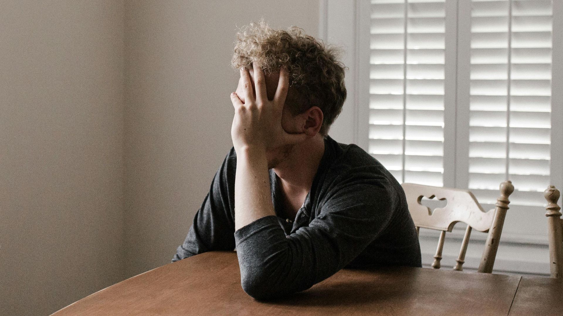 A man sits alone at a table in a bright room, displaying deep contemplation.