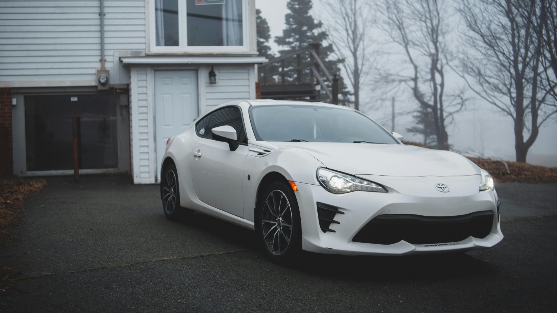 White sports car parked on driveway beside a modern house with foggy forest background.